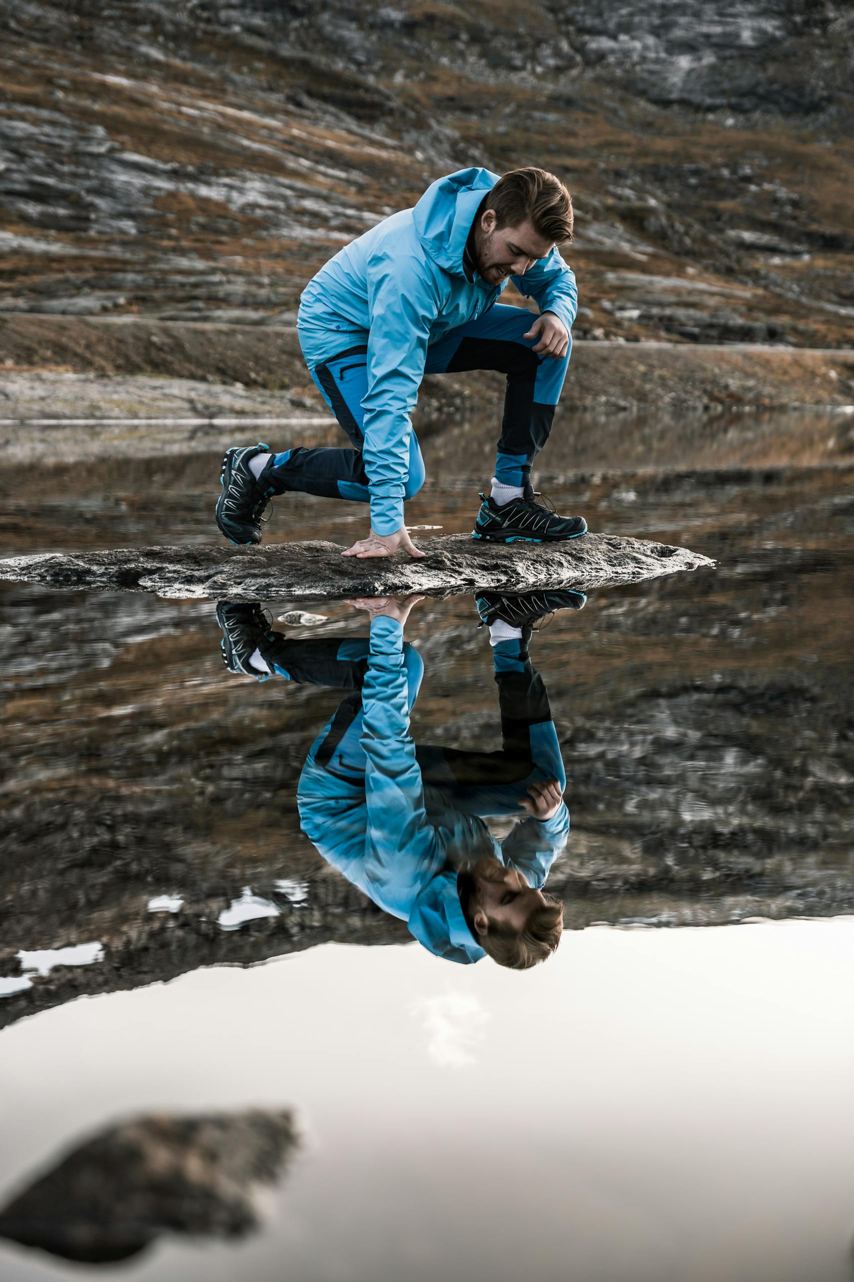 Man in blue sportswear crouching near a serene mountain lake reflection.